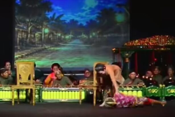 Onstage, a dancer lies on the floor beside a standing partner while a live band sits in the background.