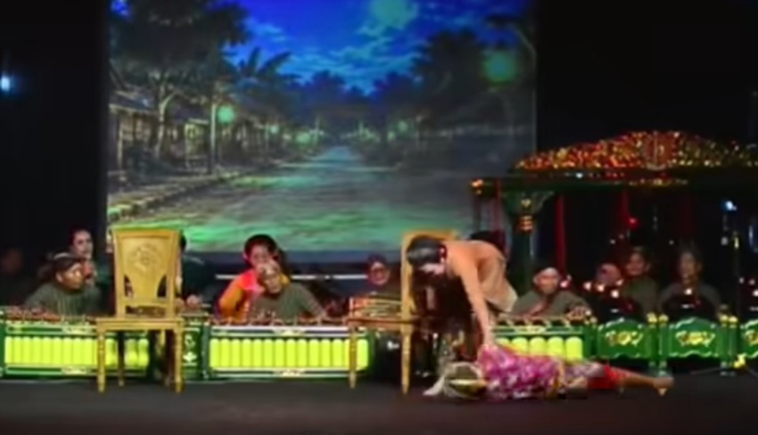 Onstage, a dancer lies on the floor beside a standing partner while a live band sits in the background.