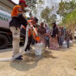 Volunteers in orange vests fill plastic jugs with water from a tanker while villagers queue along a dirt road. A banner on the truck reads 'Air Bersih Baznas'.