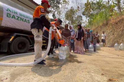 Volunteers in orange vests fill plastic jugs with water from a tanker while villagers queue along a dirt road. A banner on the truck reads 'Air Bersih Baznas'.