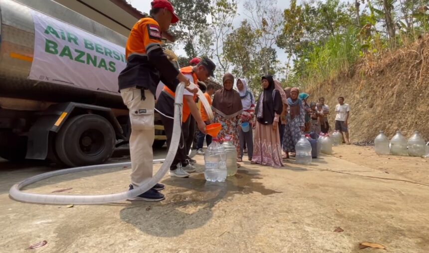 Volunteers in orange vests fill plastic jugs with water from a tanker while villagers queue along a dirt road. A banner on the truck reads 'Air Bersih Baznas'.