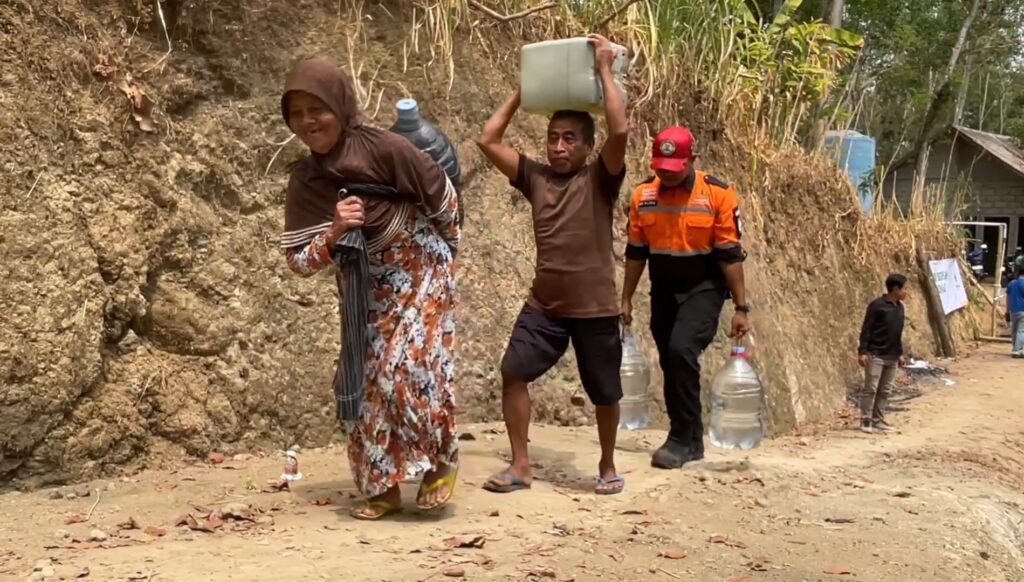 People carrying large water containers along a dusty hillside path; a woman in a floral dress leads, followed by men with jugs and a worker in orange uniform