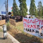 Group of people protesting along a rural road with banners and signs under a blue sky.