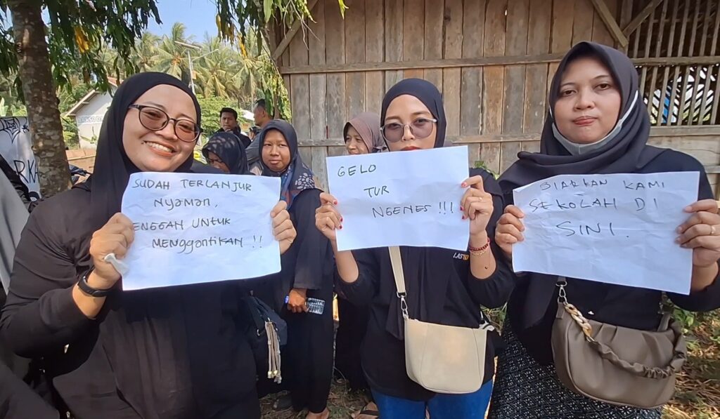 Group of women wearing hijabs hold handwritten protest signs outdoors beside a wooden wall.