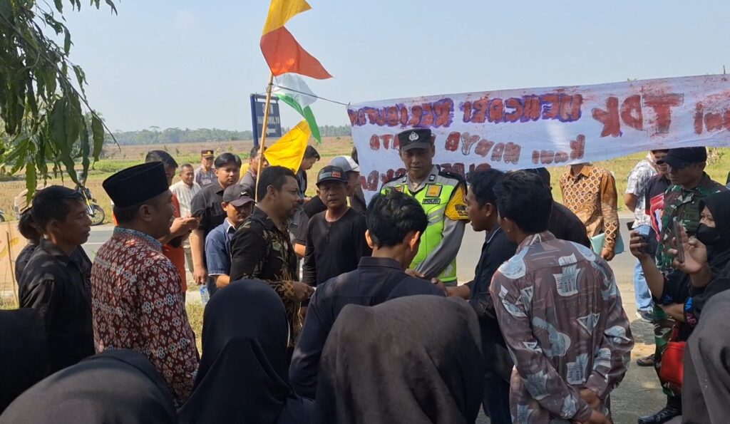 Crowd of people gathered on a roadside with a police officer in a bright vest and a banner in the background. Flags flutter above them on a sunny day.