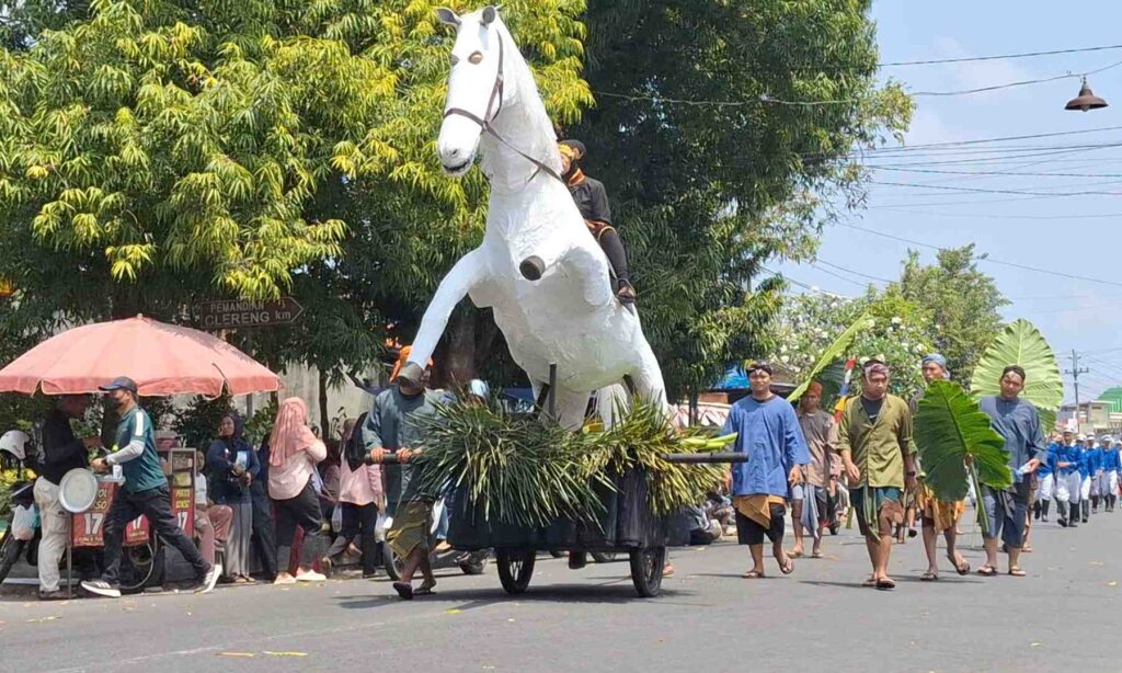 A large white horse sculpture on a wheeled cart being paraded down a street, with spectators and participants along the roadside.