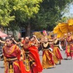 Dancers in red and gold costumes perform a street parade, followed by a large yellow phoenix float with golden decorations in the background.