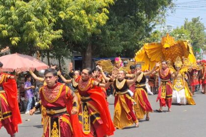 Dancers in red and gold costumes perform a street parade, followed by a large yellow phoenix float with golden decorations in the background.