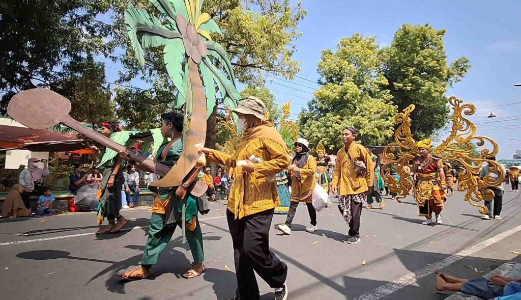 Group of performers in yellow and green costumes marching in a sunny street parade, carrying oversized decorative props and a large palm-tree structure.