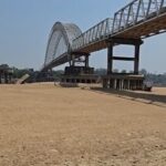 Steel arch/Truss bridge spanning a dry, sandy riverbed with concrete supports in the distance