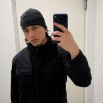 Young man in a black beanie and jacket taking a mirror selfie with a dark smartphone.