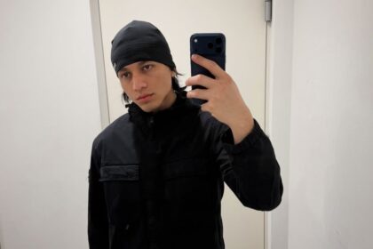 Young man in a black beanie and jacket taking a mirror selfie with a dark smartphone.