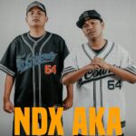 Two men in matching baseball-style jerseys stand side by side, one in black and the other in white, with bold orange text 'NDX AKA' across the bottom.