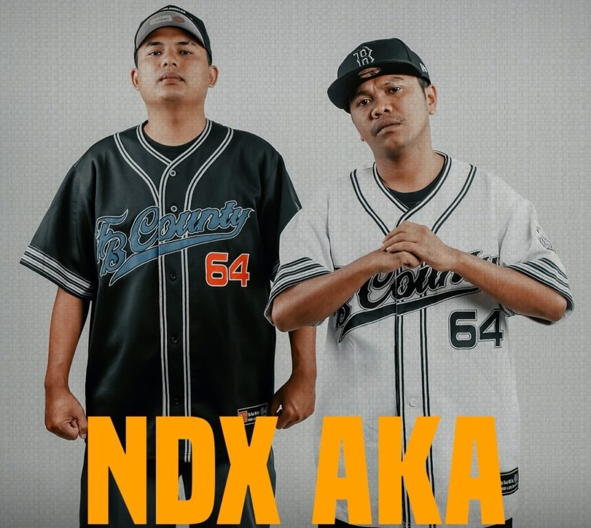 Two men in matching baseball-style jerseys stand side by side, one in black and the other in white, with bold orange text 'NDX AKA' across the bottom.