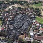 Aerial view of a village with a large circular burn area in the center and surrounding intact houses and vegetation.