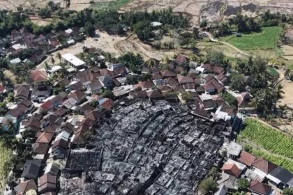 Aerial view of a village with a large circular burn area in the center and surrounding intact houses and vegetation.