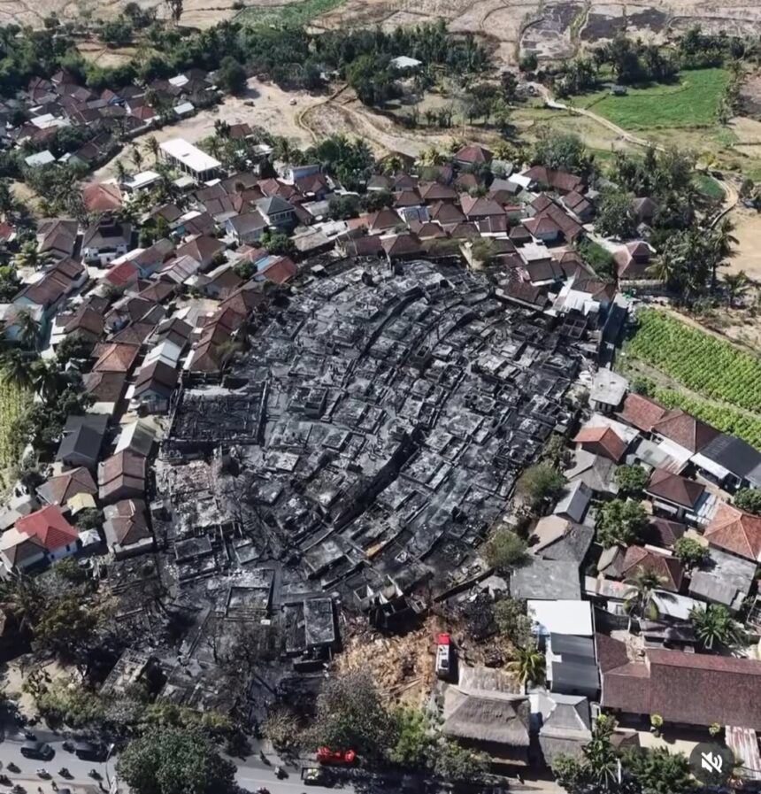 Aerial view of a village with a large circular burn area in the center and surrounding intact houses and vegetation.