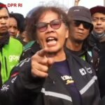 Man wearing sunglasses and a dark jacket shouts and points at the camera during a street protest, surrounded by a crowd of reporters and bystanders.