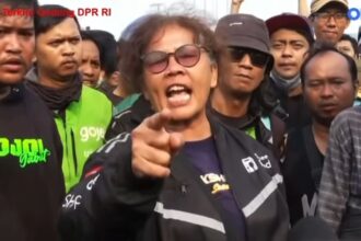 Man wearing sunglasses and a dark jacket shouts and points at the camera during a street protest, surrounded by a crowd of reporters and bystanders.