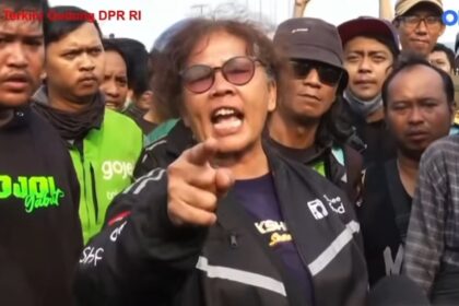 Man wearing sunglasses and a dark jacket shouts and points at the camera during a street protest, surrounded by a crowd of reporters and bystanders.