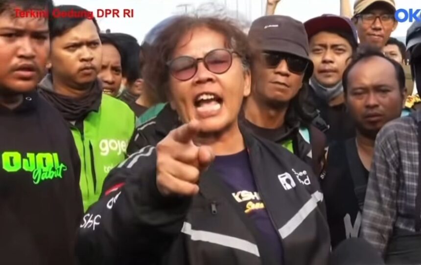 Man wearing sunglasses and a dark jacket shouts and points at the camera during a street protest, surrounded by a crowd of reporters and bystanders.