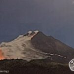 Night view of a volcano erupting with lava glowing at the base and smoke rising from the summit; logos from Kementerian ESDM and Badan Geologi visible in corners.