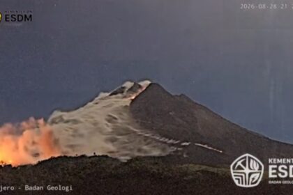 Night view of a volcano erupting with lava glowing at the base and smoke rising from the summit; logos from Kementerian ESDM and Badan Geologi visible in corners.