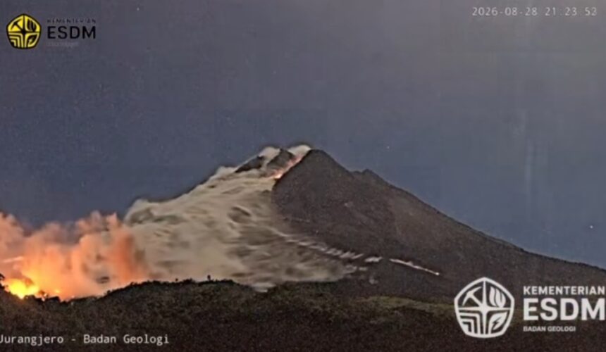 Night view of a volcano erupting with lava glowing at the base and smoke rising from the summit; logos from Kementerian ESDM and Badan Geologi visible in corners.