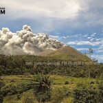 Volcano erupting with a tall smoke plume over a forested hillside; logo and watermark from Indonesian Geology Agency in the top-left corner.