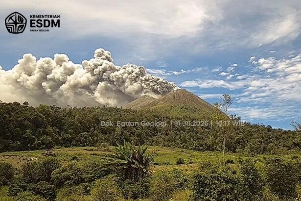 Volcano erupting with a tall smoke plume over a forested hillside; logo and watermark from Indonesian Geology Agency in the top-left corner.