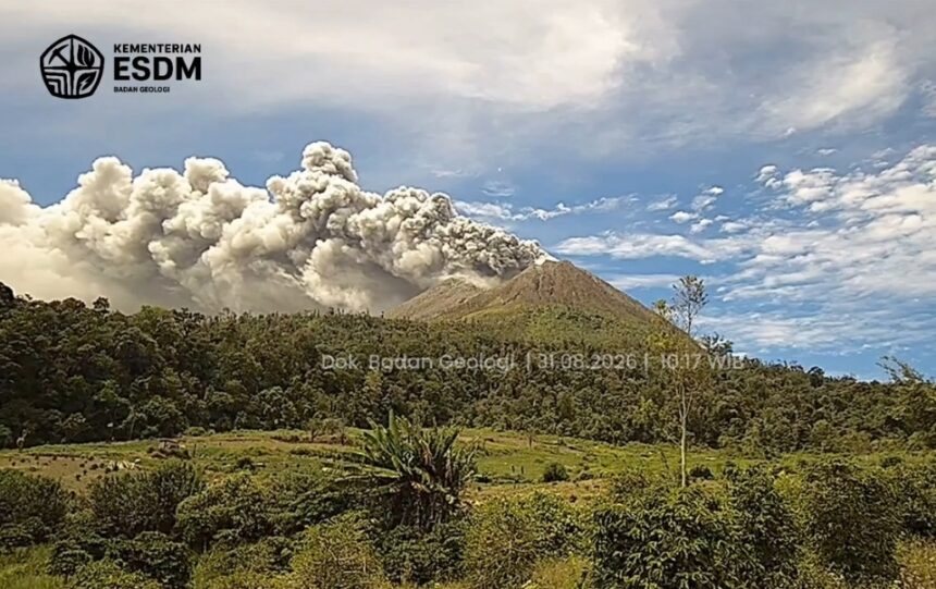 Volcano erupting with a tall smoke plume over a forested hillside; logo and watermark from Indonesian Geology Agency in the top-left corner.