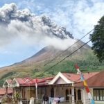Volcano erupting with a tall gray plume rising over a village, foreground shows houses with red roofs and power lines.