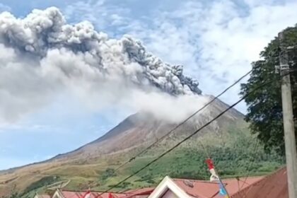 Volcano erupting with a tall gray plume rising over a village, foreground shows houses with red roofs and power lines.