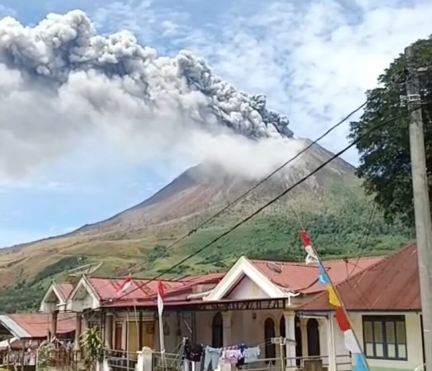 Volcano erupting with a tall gray plume rising over a village, foreground shows houses with red roofs and power lines.