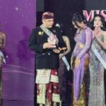 Man in traditional Indonesian attire receives a trophy on stage, flanked by contestants in glittering evening gowns at a Miss pageant event.
