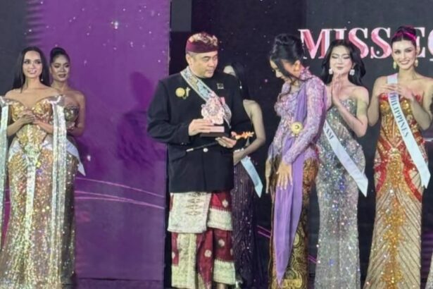 Man in traditional Indonesian attire receives a trophy on stage, flanked by contestants in glittering evening gowns at a Miss pageant event.