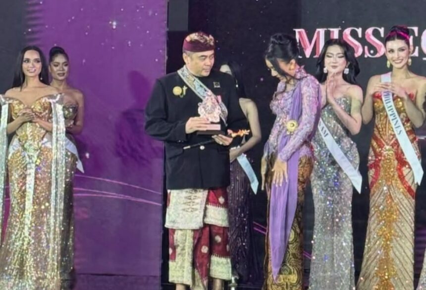 Man in traditional Indonesian attire receives a trophy on stage, flanked by contestants in glittering evening gowns at a Miss pageant event.