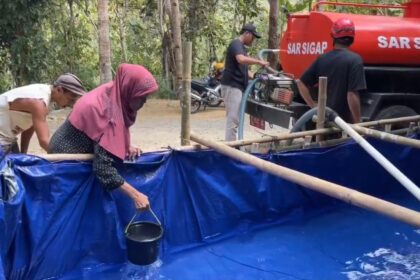 Group of villagers near a blue plastic-lined water tank, filling buckets as a red tanker labeled 'SAR SIGAP' stands nearby.