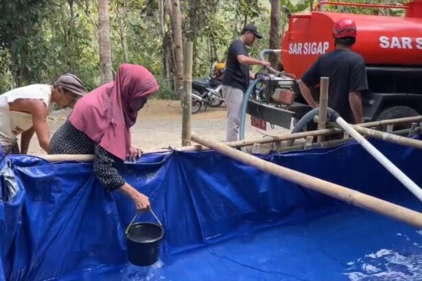 Group of villagers near a blue plastic-lined water tank, filling buckets as a red tanker labeled 'SAR SIGAP' stands nearby.