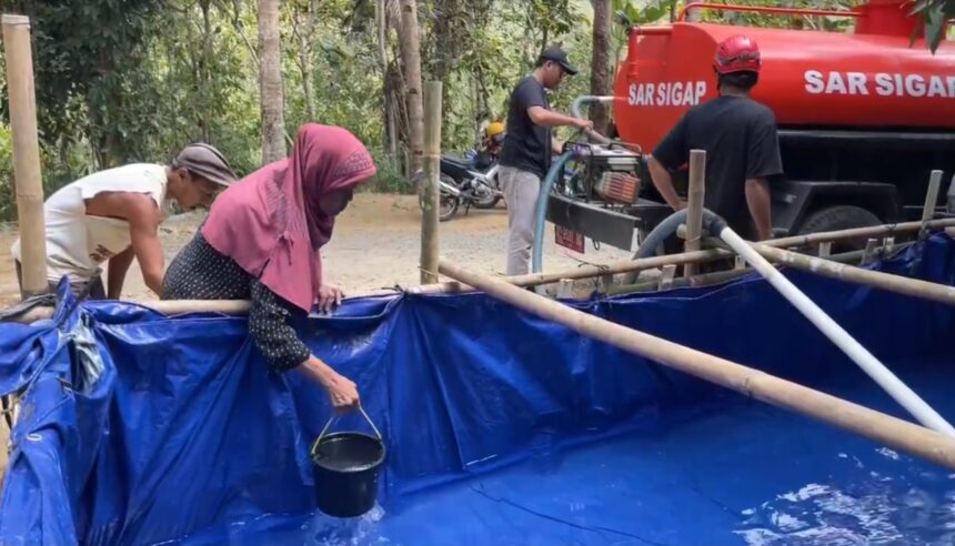 Group of villagers near a blue plastic-lined water tank, filling buckets as a red tanker labeled 'SAR SIGAP' stands nearby.