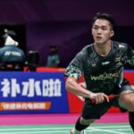 Male badminton player in a black patterned jersey lunges to hit a shuttlecock on a colorfully lit court with sponsor banners in the background, during a match.