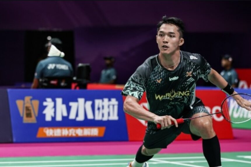Male badminton player in a black patterned jersey lunges to hit a shuttlecock on a colorfully lit court with sponsor banners in the background, during a match.