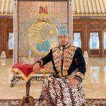 Man in ornate ceremonial outfit with a blue hat sits beside a small table covered with a red cloth inside a grand hall, with a decorative backdrop behind him.