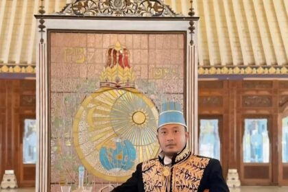 Man in ornate ceremonial outfit with a blue hat sits beside a small table covered with a red cloth inside a grand hall, with a decorative backdrop behind him.