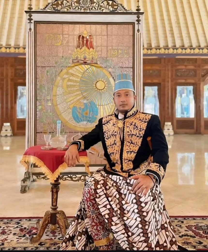 Man in ornate ceremonial outfit with a blue hat sits beside a small table covered with a red cloth inside a grand hall, with a decorative backdrop behind him.