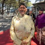 Man in pale gold floral baju Melayu with a black turban stands on a red carpet at an outdoor ceremony; others in traditional dress in the background.