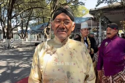 Man in pale gold floral baju Melayu with a black turban stands on a red carpet at an outdoor ceremony; others in traditional dress in the background.
