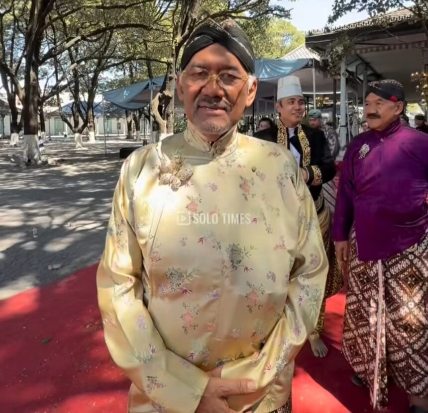 Man in pale gold floral baju Melayu with a black turban stands on a red carpet at an outdoor ceremony; others in traditional dress in the background.