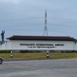 Entrance sign for Yogyakarta International Airport with a statue on the left and a motorcyclist on the road in the foreground.