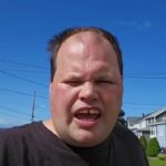 Man in a black shirt speaks to the camera outdoors on a sunny day, with houses and a blue sky in the background.